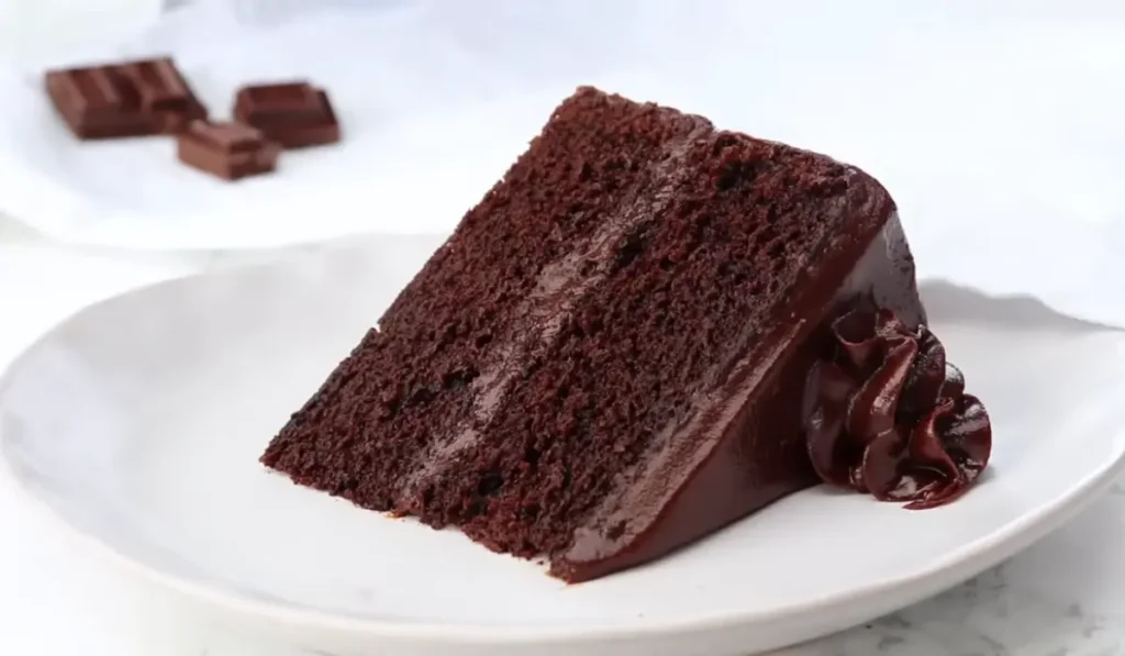 "Slice of moist chocolate cake topped with smooth chocolate frosting on a white plate."